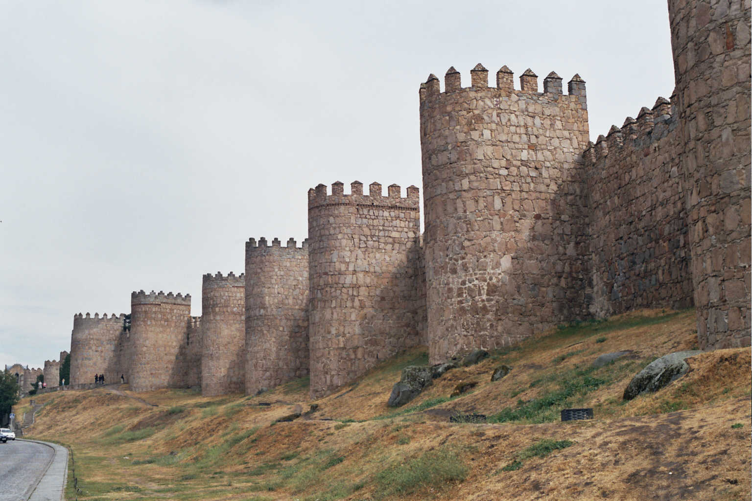 Cubos de la muralla de Ávila sobre un talud de hierba seca, carretera al pie.